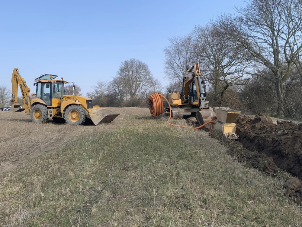 Gravemaskine med orange slange, der udfører dræningsarbejde på en mark