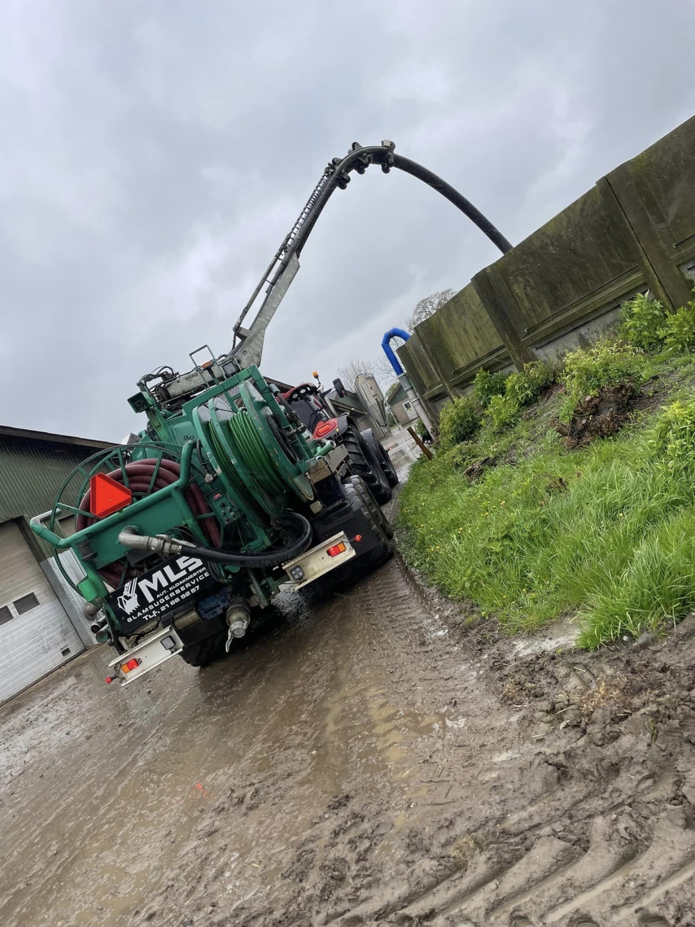 Kloakspulingsmaskine i gang med spuling ved en gård, med sugeslangen over muren.